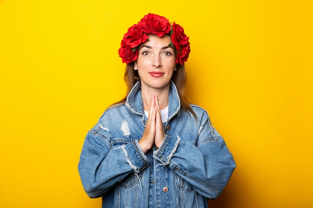Jeune femme dans une veste en jean et une couronne de fleurs rouges sur sa tête croisa les bras dans une pose de yoga sur un mur jaune.