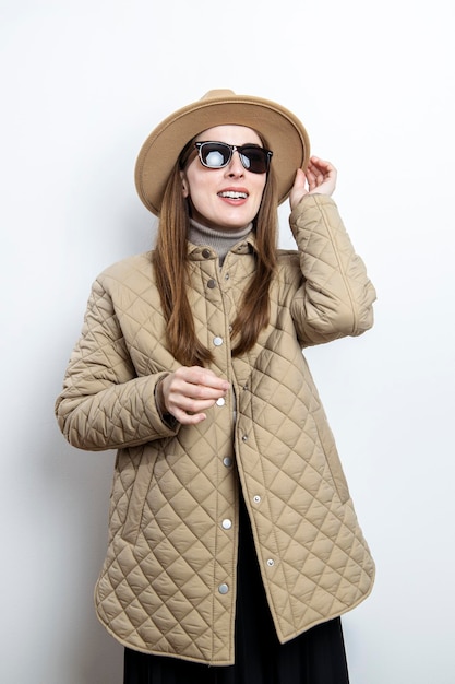 Photo jeune femme dans une veste en lunettes de soleil dans un chapeau lève les yeux sur le fond d'un mur blanc