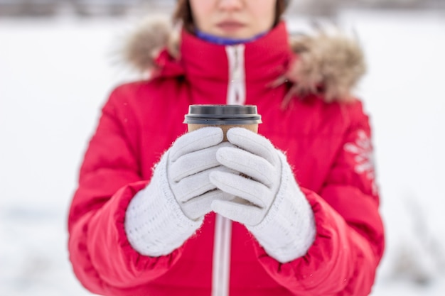 Une jeune femme dans une veste rouge en hiver tient un verre de café ou de thé chaud