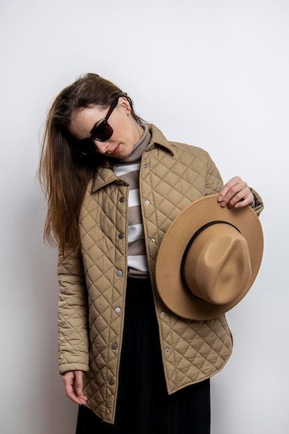 Jeune femme dans une veste tenant un chapeau regardant vers le bas contre un mur blanc