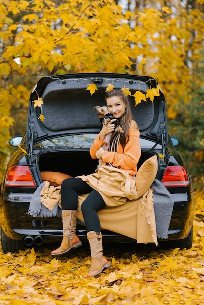 Une jeune femme dans une voiture sur le coffre dans le contexte de la forêt d'automne est assise détendue et embrasse son chien préféré