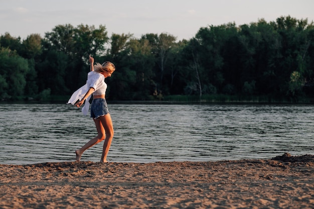 Une jeune femme danse au bord de la rivière une belle blonde heureuse en chemise blanche et short en jean...