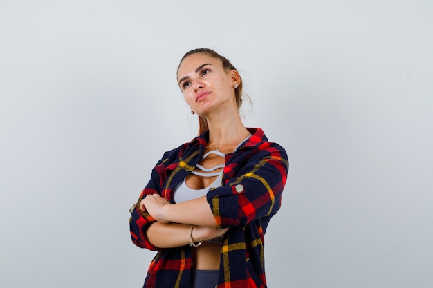 Jeune femme debout avec les bras croisés en haut, chemise à carreaux et regardant pensive, vue de face.