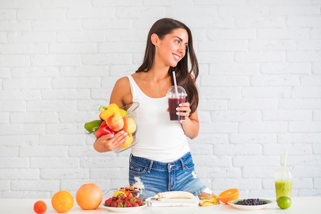 Photo jeune femme, debout, contre, mur, tenue, bol, de, légumes frais, et, fruits, et, jus