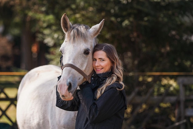 Une jeune femme debout à côté d'un cheval arabe blanc, des arbres floues, un fond en gros plan.