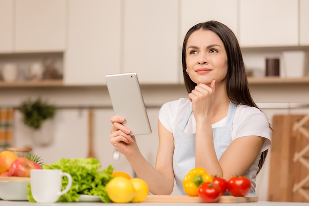 Jeune femme debout dans la cuisine avec tablette, ayant une idée et à la recherche de recettes dans la cuisine. Vue de côté