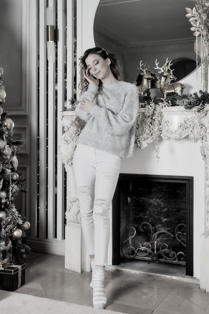 Jeune femme debout devant la cheminée et l'arbre de Noël avec des cadeaux heureux et rêvant. Couleur argent blanc sur fond de salon pour joyeux Noël et bonne année. Moments d'ambiance en famille