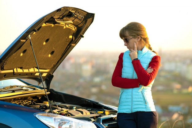 Jeune femme debout près de voiture en panne avec capot sauté ayant des problèmes avec son véhicule.