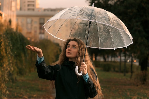 Jeune femme debout sous un parapluie transparent regarde pour voir s'il pleut Fille aux cheveux bruns se promène dans le parc d'automne