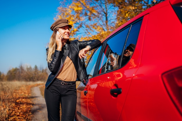 Jeune femme debout en voiture à l'aide de smartphone sur la route de l'automne. Pilote arrêté auto en forêt pour appeler