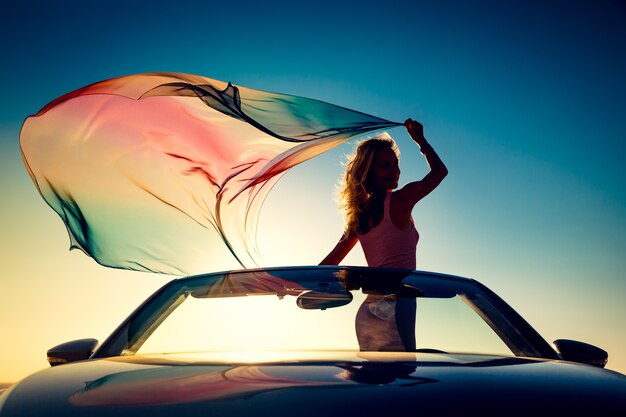 Jeune femme debout sur une voiture avec un tissu volant sur fond de ciel coucher de soleil