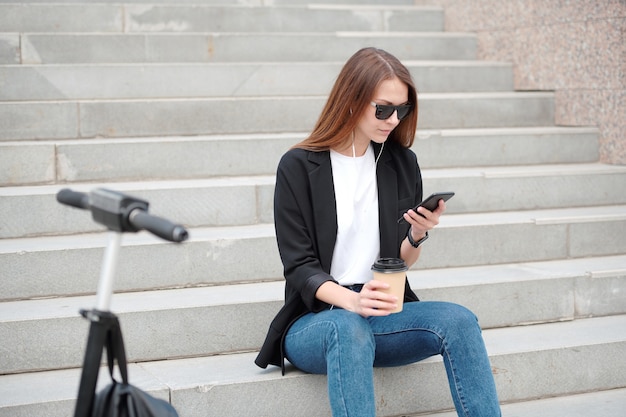 Jeune femme décontractée aux cheveux longs assis sur l'escalier en milieu urbain et défilement dans le smartphone tout en écoutant de la musique