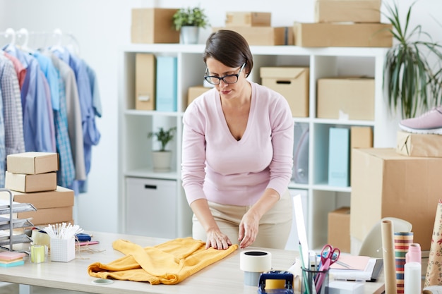 Jeune femme décontractée se penchant sur le bureau tout en pliant le pull en coton