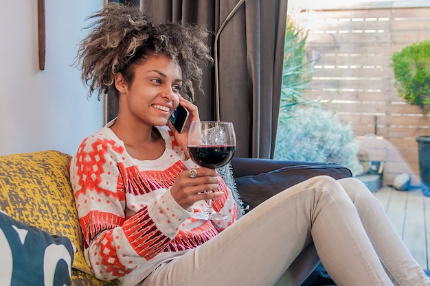 Une jeune femme décontractée utilisant un téléphone portable assis sur son divan chez soi et jouit d&#39;un verre de vin. Africaine, Américain, femme, conversation, téléphone portable