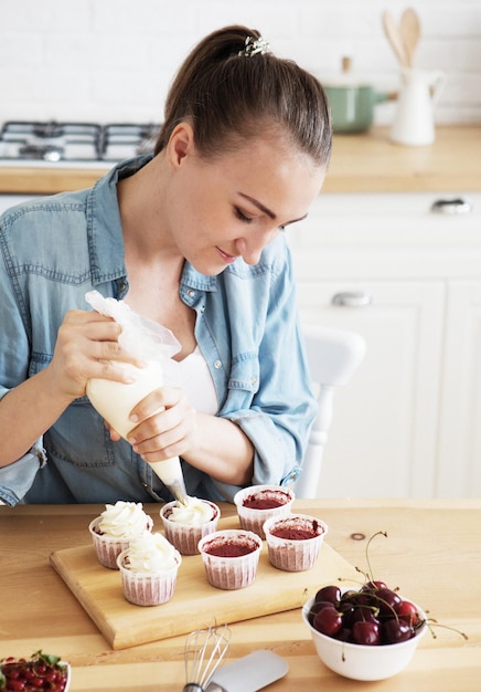 Une jeune femme décore des muffins à la crème blanche dans sa cuisine