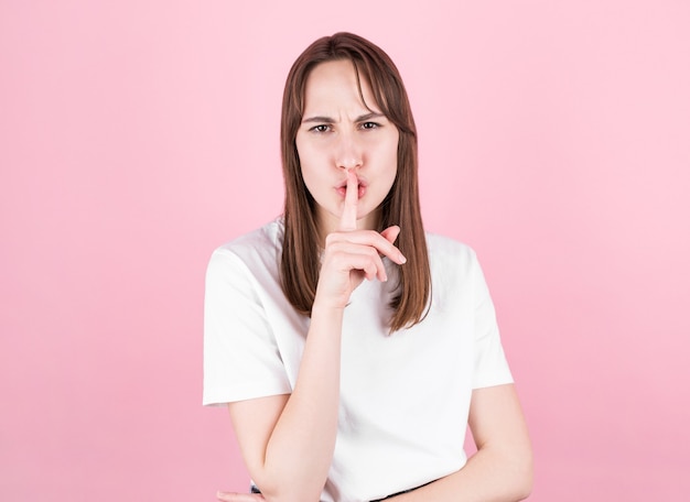 Jeune femme demandant à être calme avec le doigt sur les lèvres. Silence et concept secret sur fond rose,