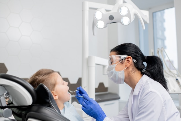 Jeune femme dentiste en blanchon, lunettes de protection, masque et gants va faire un bilan oral de la cavité buccale de son petit patient