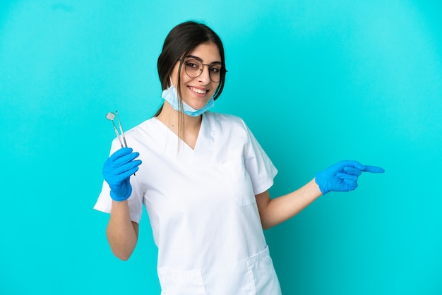 Jeune femme dentiste de race blanche tenant des outils isolés sur fond bleu, pointant le doigt sur le côté