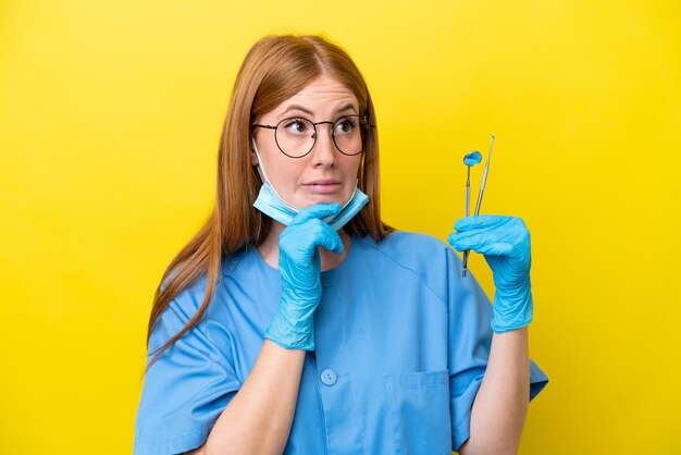 Jeune femme dentiste rousse isolée sur fond jaune ayant des doutes et pensant