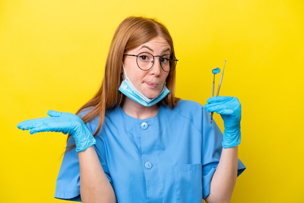 Jeune femme dentiste rousse isolée sur fond jaune ayant des doutes tout en levant les mains