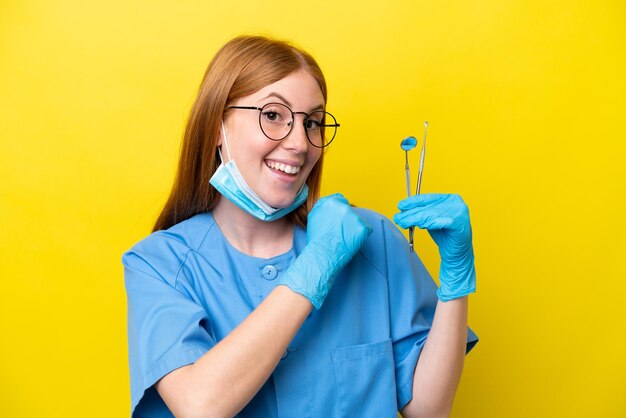 Jeune femme dentiste rousse isolée sur fond jaune célébrant une victoire