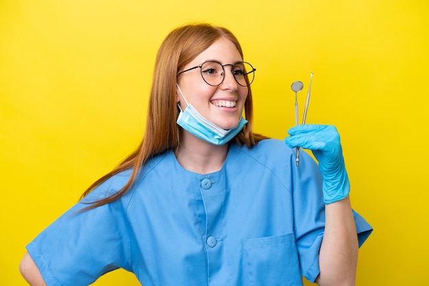 Jeune femme dentiste rousse isolée sur fond jaune posant avec les bras à la hanche et souriant