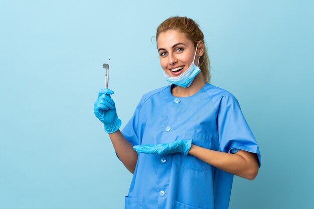 Jeune femme dentiste tenant des outils sur bleu isolé s'étendant les mains sur le côté pour inviter à venir