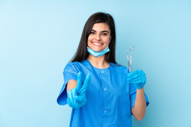 Jeune femme dentiste tenant des outils sur le mur bleu isolé souriant et montrant le signe de la victoire