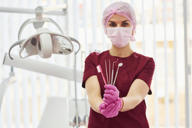 Jeune femme dentiste en uniforme debout dans le bureau de stomatologie tenant une brosse et des outils