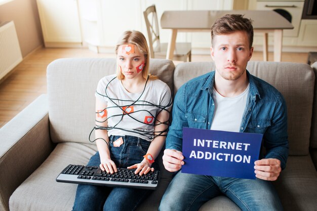 Une jeune femme a une dépendance aux médias sociaux. Assis sur le canapé sans émotion. Cordon de cou enveloppé. Zomby Internet. Un gars inquiet regarde la caméra et tient du papier sur la dépendance à Internet.