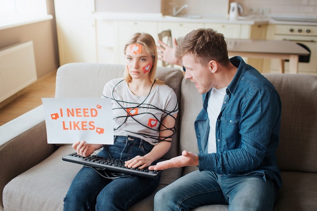 Une jeune femme a une dépendance aux médias sociaux. Assis sur le canapé sans émotion. Corps enveloppé de cordon. Mains sur le clavier. Jeune homme en colère criant. Personnes émotionnelles.