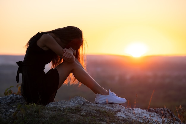 Jeune femme déprimée en robe d'été courte noire assise sur un rocher pensant à l'extérieur au coucher du soleil. Femme à la mode contemplant dans une soirée chaude dans la nature.