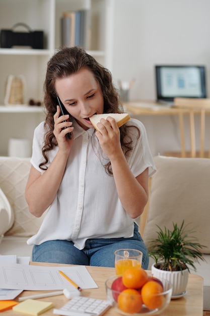 Jeune femme designer d'interface utilisateur indépendante mangeant un sandwich et parlant au téléphone avec le client