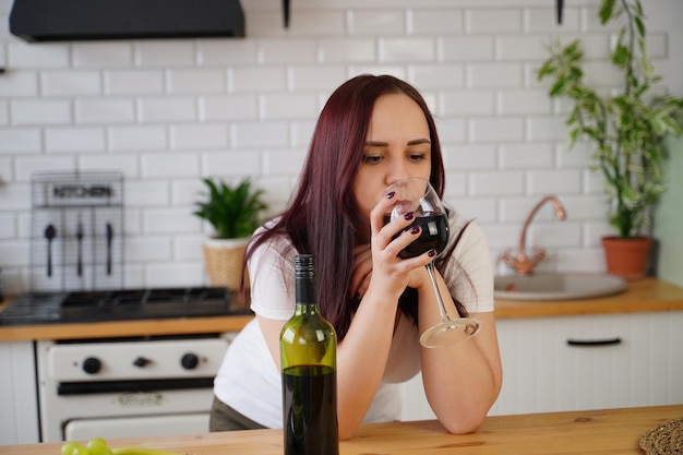 Une jeune femme détendue boit du vin rouge debout à la table de la cuisine Brune adulte se reposant avec de l'alcool dans la cuisine