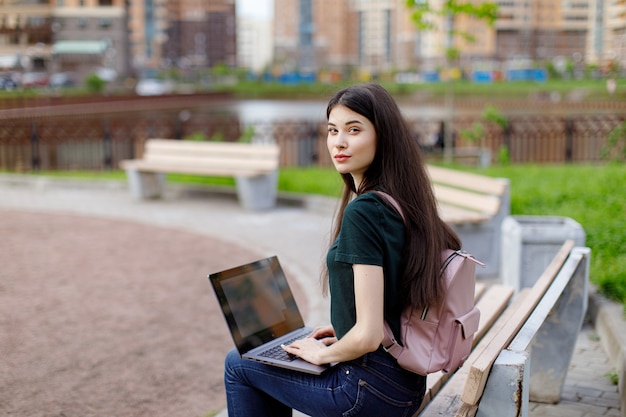 Jeune femme détendue dans un T-shirt vert et avec un sac à dos assis sur un banc en bois, boire du café et parcourir son ordinateur portable