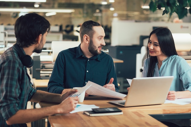 Jeune femme et deux hommes assis à la table du bureau avec des documents et un ordinateur portable et discutant de leur projet