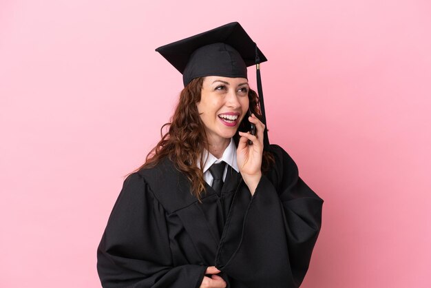 Jeune femme diplômée universitaire isolée sur fond rose en gardant une conversation avec le téléphone mobile