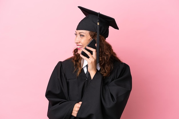 Jeune femme diplômée universitaire isolée sur fond rose en gardant une conversation avec le téléphone portable avec quelqu'un