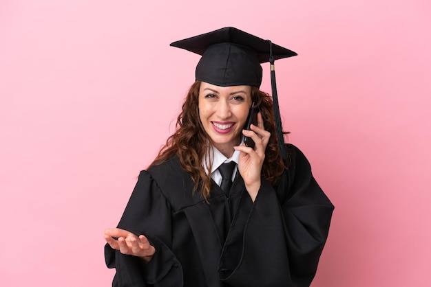 Jeune femme diplômée universitaire isolée sur fond rose en gardant une conversation avec le téléphone portable avec quelqu'un