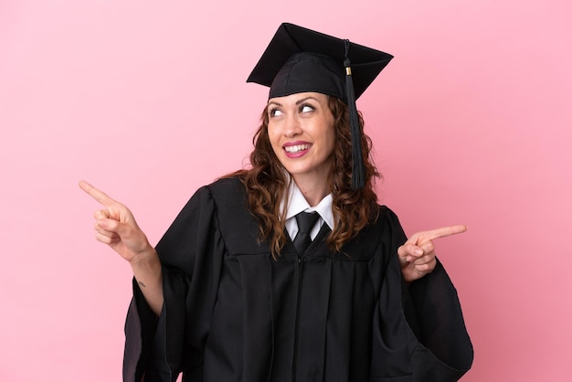 Jeune femme diplômée universitaire isolée sur fond rose pointant le doigt vers les latéraux et heureuse