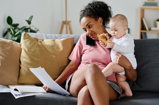 Jeune femme discutant de documents au téléphone alors qu'elle était assise sur un canapé avec son bébé