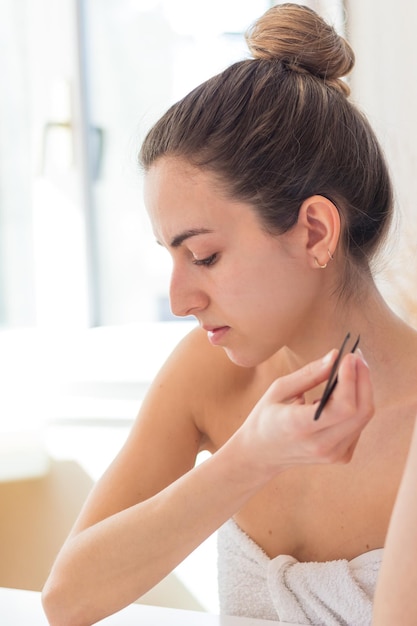 Jeune femme de douche avec chignon et serviette et pince à épiler à la main regardant vers le bas