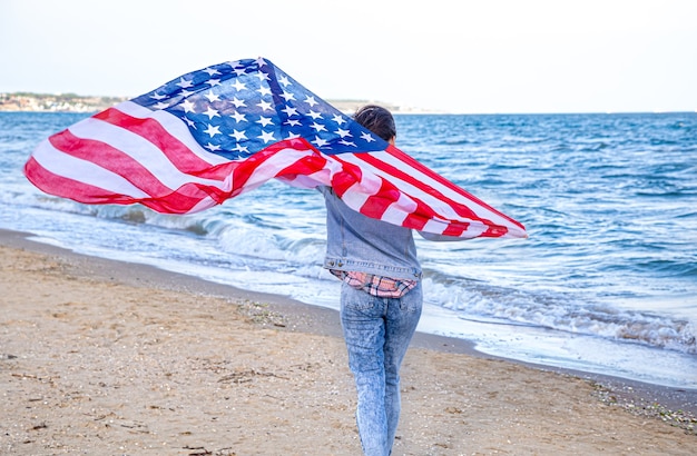 Photo une jeune femme avec un drapeau américain court au bord de la mer. le concept du patriotisme et des célébrations de la fête de l'indépendance.