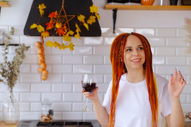 Jeune femme avec des dreadlocks lumineux buvant du vin rouge dansant dans la cuisine Portrait de femme adulte divertissant avec un verre de vin à la maison