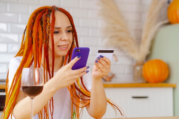 Jeune femme avec des dreadlocks lumineux avec carte de crédit de téléphone portable assis dans la cuisine Bonne femme naviguant sur smartphone entrant les détails de la carte payant l'achat sur Internet Concept de magasinage en ligne