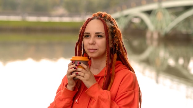 Jeune femme avec des dreadlocks tenant une tasse de café debout près du lac de la rivière sur fond de grand pont dans le parc de la ville