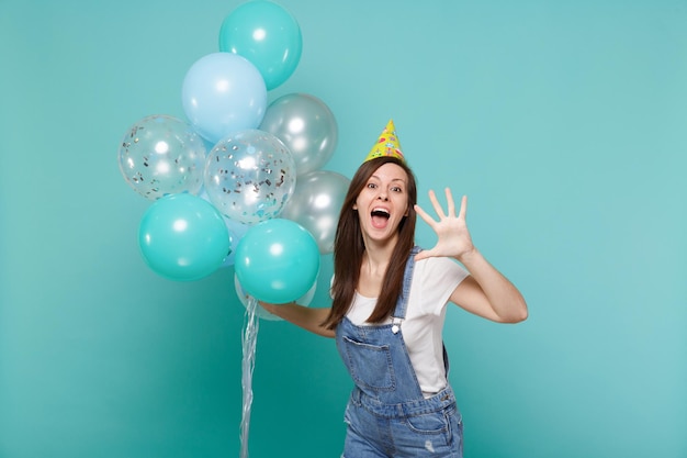 Jeune femme drôle au chapeau d'anniversaire gardant la bouche ouverte montrant la paume et célébrant, tenant des ballons à air colorés isolés sur fond bleu turquoise. Fête d'anniversaire, concept d'émotions de personnes.