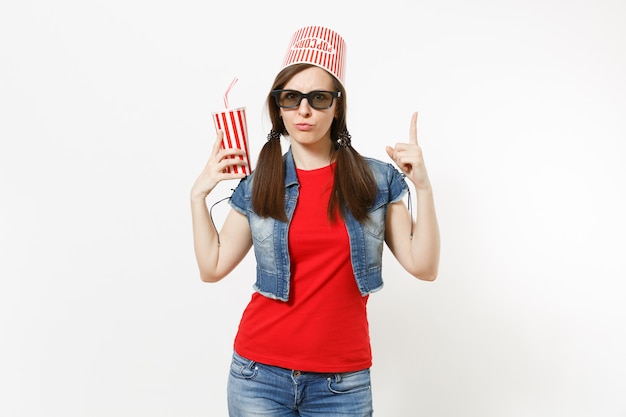 Jeune femme drôle dans des lunettes 3d avec seau pour pop-corn sur la tête en regardant un film tenant une tasse de soda, cola pointant les doigts vers le haut sur l'espace de copie isolé sur fond blanc. Les émotions au cinéma.