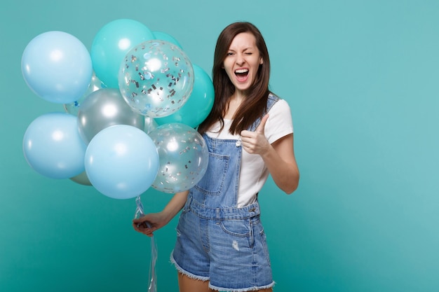 Jeune femme drôle dans des vêtements en denim clignotant montrant le pouce vers le haut, célébrant et tenant des ballons à air colorés isolés sur fond de mur bleu turquoise. Fête d'anniversaire, concept d'émotions de personnes.