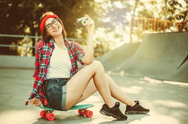 Une jeune femme drôle dans des vêtements élégants est assise sur une planche à roulettes et utilise un appareil photo rétro dans un skatepark par une belle journée ensoleillée. Notion de jeunesse